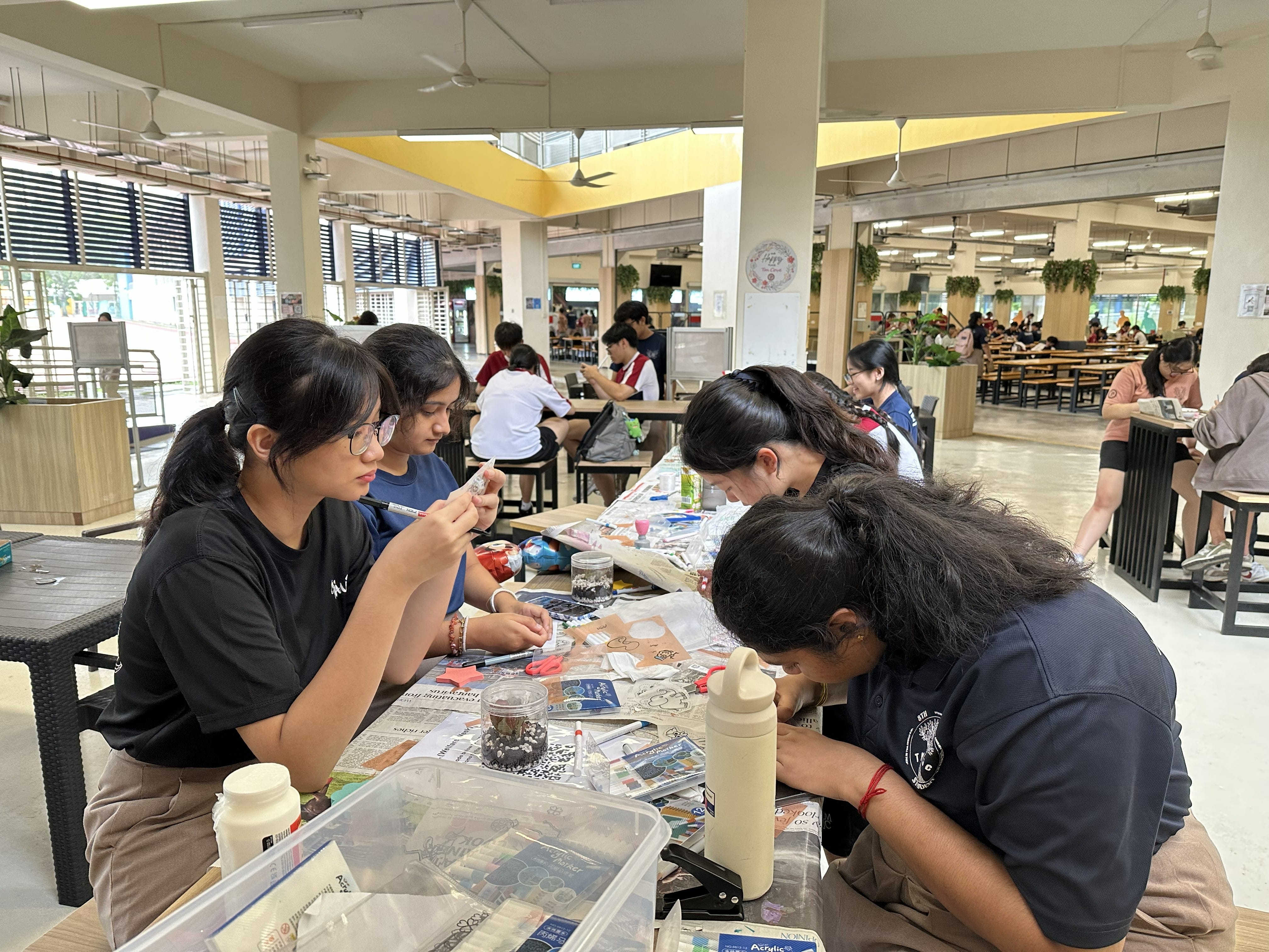 Four female students sit around a table covered in craft materials, focused on designing and cutting shrink plastic keychains in a school canteen setting.